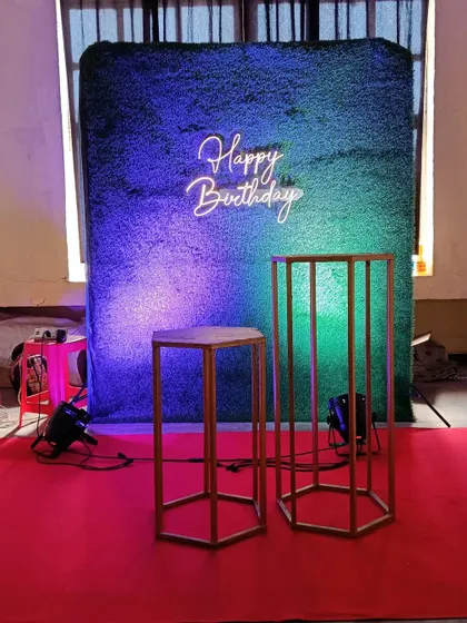 A minimalist yet effective birthday setup, featuring a green turf wall, a 'Happy Birthday' neon sign, and display pedestals, ready for a cake or other decor.