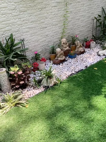 This garden border combines a lush artificial lawn with a wide band of white pebbles, which serves as a clean backdrop for a collection of monk statues and other decorative planters.