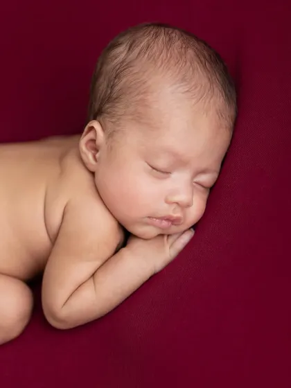 A simple, powerful portrait of a newborn sleeping on their side against a deep red backdrop. This image captures the stillness and calm of the very beginning.