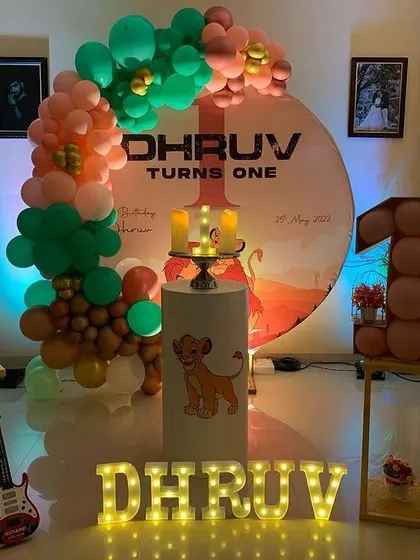 A Lion King themed first birthday party setup at home. This decoration features a custom circular backdrop, a balloon garland, a cake stand with a Simba graphic, and light-up letters spelling the name.