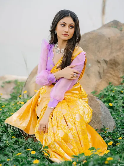 Sanjana Rao in a thoughtful pose, surrounded by yellow flowers that match her beautiful saree. The puff sleeves of the blouse add a modern, romantic touch.