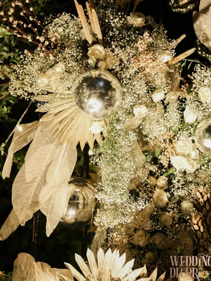 A detailed shot of a nighttime floral arrangement. The combination of baby's breath, white roses, dried elements, and a silver disco ball creates a fascinating mix of textures that comes alive under the lights.