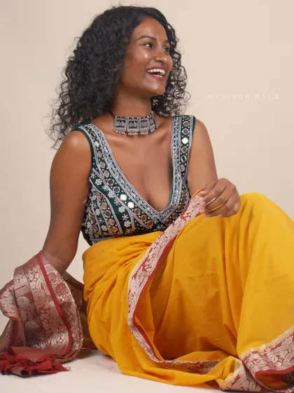A bright and cheerful portrait combining the deep green mirror-work blouse with a vibrant yellow saree. The model's joyful expression and relaxed pose create a warm and inviting image that celebrates color and traditional style.