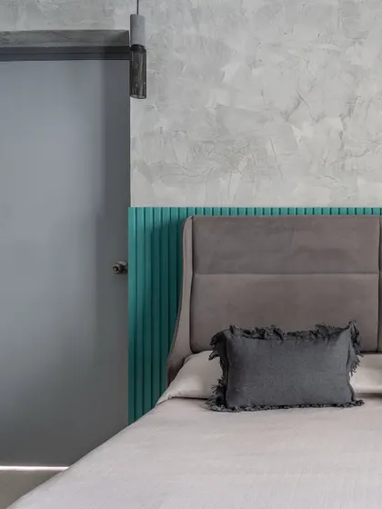 A detail of the guest bedroom, showing the contrast between the grey upholstered headboard, teal fluted panel, and the concrete-textured wall.