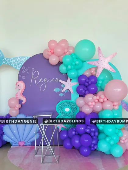 A purple and teal mermaid theme with a large shell cutout and mermaid tail accents. The balloon garland and seahorse details create a beautiful underwater photo corner.