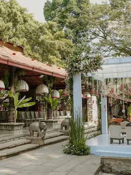 A wider perspective of the wedding setup, showing the mandap, the surrounding traditional architecture, and the guest seating under large, floral-adorned woven lampshades.