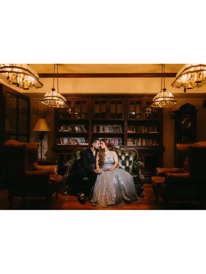 An elegant couple portrait in a vintage-style library, sharing a kiss on a classic leather sofa under dramatic lighting.
