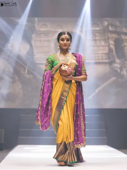 This model walks the runway in a beautiful yellow and purple saree. The soft lighting from above highlights the rich colors and the traditional elegance of her attire.