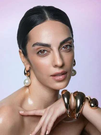 A close-up beauty shot showcasing luminous skin and bold, natural brows. The slicked-back hair and statement jewelry complement the clean makeup perfectly.