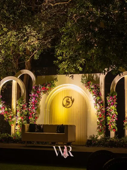 A wider view of the romantic white arched stage, nestled amongst the trees. The soft lighting highlights the floral arrangements and the couple's monogram, creating a perfect backdrop for photos.