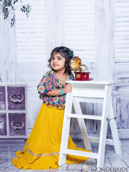 A little girl in a yellow lehenga and a colorful top, posing by a white step ladder and gramophone. The vintage-style backdrop adds a timeless feel to this child portrait.