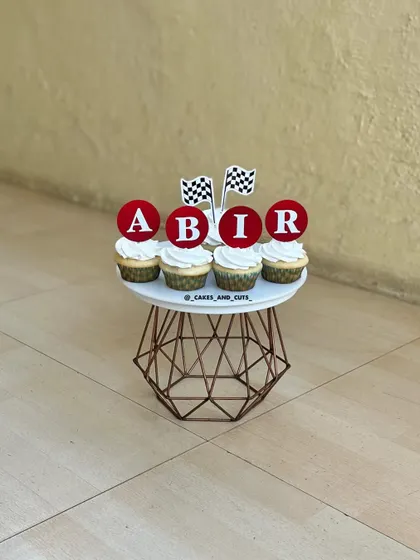 Customized cupcakes for a race car theme, featuring checkered flags and personalized "ABIR" toppers.