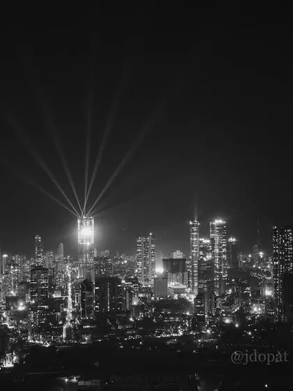 A black and white photograph of the Mumbai skyline at night during Diwali, with spotlights from the Palais Royale creating a dramatic effect.