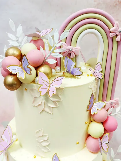 We craft an ethereal dream with this two-tiered rainbow butterfly cake. A soft pastel rainbow arch, delicate butterflies, and clusters of pink, gold, and cream spheres come together to create a whimsical and sophisticated masterpiece.