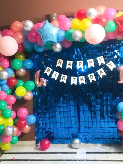 A colorful home birthday decoration for a first birthday. A blue foil curtain is framed by a multi-colored balloon arch.