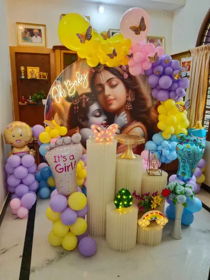 A full view of a Krishna-themed baby shower. The setup is rich with colorful balloons, themed props, and decorative plinths.