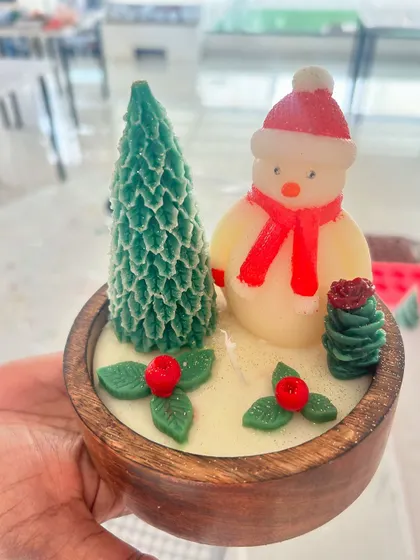 A festive Christmas-themed candle with a snowman and a Christmas tree in a wooden bowl.