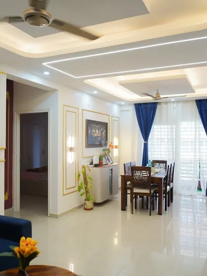 This wide view showcases a cohesive living and dining area, defined by its elegant false ceiling with recessed and profile lighting. The combination of a plush blue sofa and a classic wooden dining set creates distinct zones within the open-plan space.