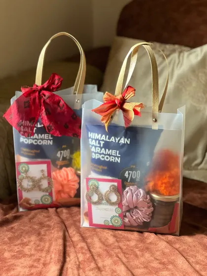 A pair of matching Rakhi gift bags for sisters, each containing gourmet popcorn, earrings, and scrunchies, tied with a festive red bow.