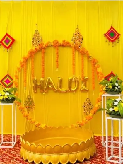 A similar version of the modern Haldi setup with a circular marigold frame, "HALDI" foil balloons, and a traditional 'urli'.