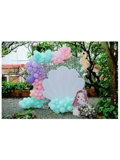 An outdoor photo-op spot for a mermaid party. A giant seashell prop is framed by a pastel balloon garland and a shimmering silver backdrop.