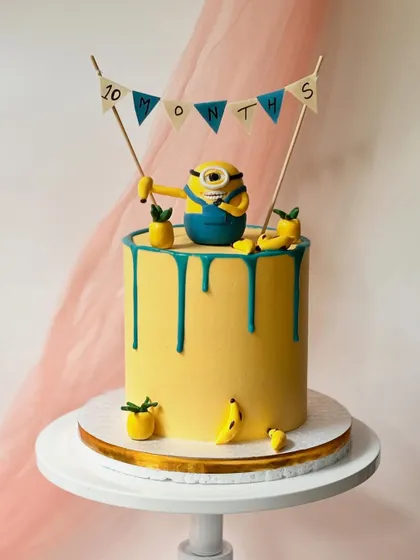 This Minion is ready to celebrate a 10-month milestone. The cake has a yellow buttercream base with a teal drip, and is decorated with fondant bananas and pineapples for a tropical feel.