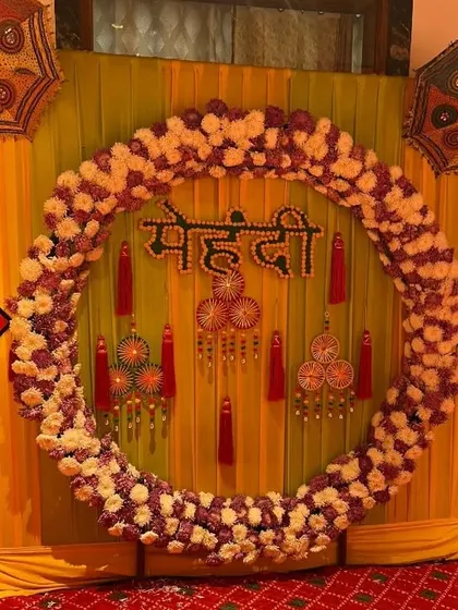 A Mehendi photo booth with a circular frame made of artificial flowers against a yellow backdrop, accented with traditional hanging ornaments.