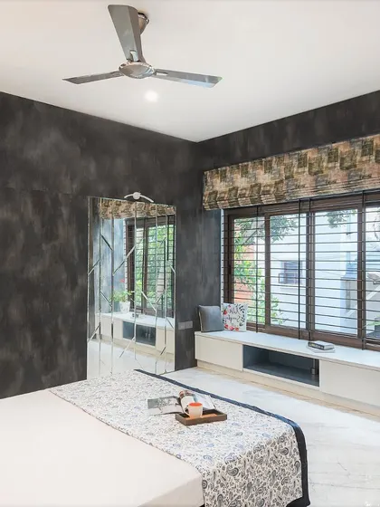 This bedroom in the Twin Courtyard House features dark, textured walls that create a cozy and intimate atmosphere. A long, low-slung window seat provides a perfect reading nook with views of the greenery outside.
