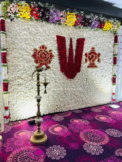 A stunning naming ceremony backdrop made of white flowers, with a central 'Namam' symbol created from red roses. A traditional brass lamp in the foreground adds a sacred glow.
