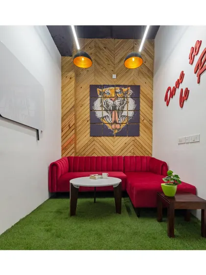 A cozy breakout nook features a bold red sofa, a tiger graphic on a chevron-patterned wood wall, and an artificial grass floor. This combination of textures and vibrant colors creates a fun and stimulating corner for relaxation.