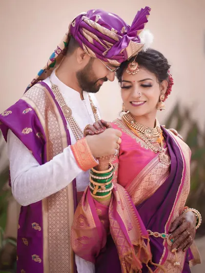 A tender moment between a bride and groom in their stunning purple and pink Maharashtrian wedding outfits. The groom gently holds her hand, creating a classic, romantic portrait.