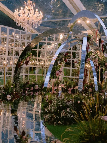 A one-of-a-kind contemporary mandap with mirrored arches, set within a massive glasshouse. This installation, surrounded by dreamy fresh florals, was designed to reflect the couple's modern personality.