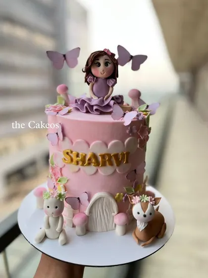 A charming fairy garden cake in a soft pink hue. The design includes a fairy topper, whimsical mushrooms, and cute woodland creatures like a bunny and a deer.