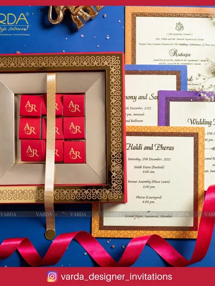 This invitation box combines tradition with a sweet touch. It features a set of personalized chocolates with the couple's initials, presented alongside a full suite of colorful wedding cards with intricate borders.