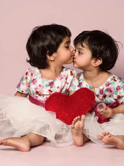 Twin sisters share a sweet kiss. This adorable photo with a red heart prop is a perfect capture of sisterly love.