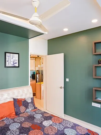 A simple yet stylish bedroom with a deep green accent wall. The custom open-shelf unit provides a lightweight and modern storage solution for books and decor.