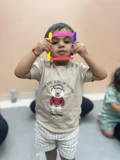 A child playfully holds up a shape made from popsicle sticks, demonstrating learning through hands-on exploration.