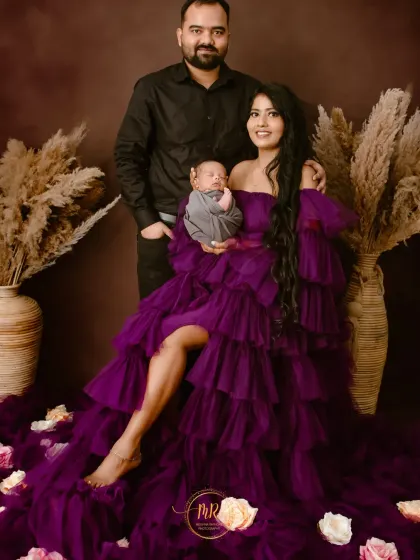 A beautiful first family portrait. The mother is seated in a stunning purple gown, holding her newborn, with her partner standing behind, creating a classic composition.