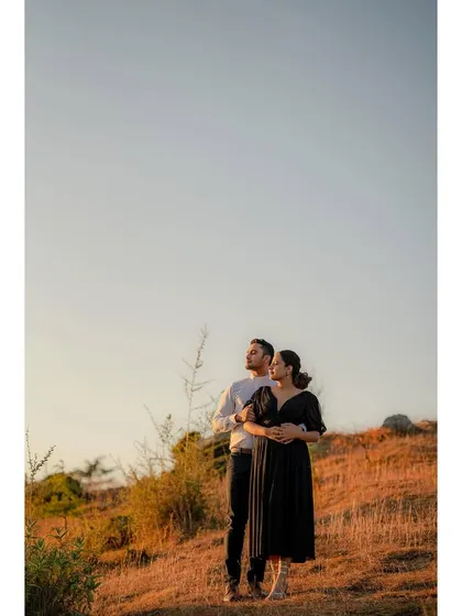 A wide shot of a couple looking out at the sunset from a hilltop. The golden light and expansive view create a serene and romantic mood.