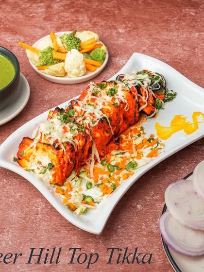 Paneer Hill Top Tikka consists of cubes of paneer marinated in a spicy red masala, grilled in the tandoor, and then topped with grated cheese. It's served with mint chutney and a side of fresh salad.