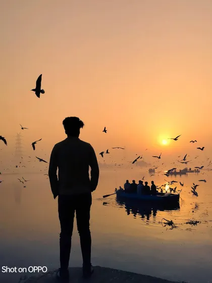 A silhouette of a person watching the sunrise over the Yamuna Ghat, as birds fly past and a boat full of people enjoys the view. It's a classic, beautiful morning scene.