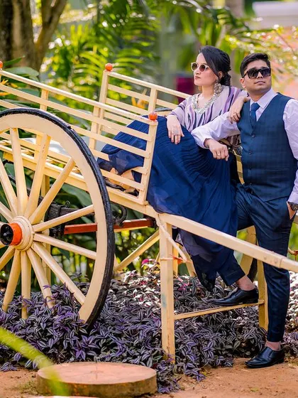 A stylish, editorial-style portrait with a rustic bullock cart. The couple's modern outfits and confident poses create a cool contrast with the traditional prop.