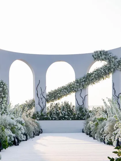An architectural statement for a seaside ceremony. This modern, curved mandap is adorned with a sweeping garland of white flowers and delicate branches, creating a clean, elegant, and artistic focal point against the open sky.