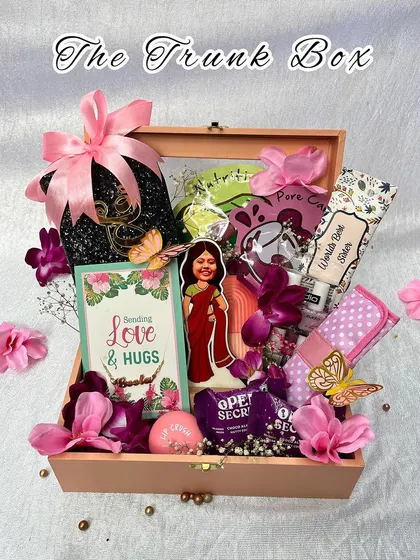 "The Trunk Box" is a beautiful wooden chest filled with a curated selection of gifts for her. It includes snacks, skincare, a personalized caricature, and other thoughtful items, decorated with pink flowers.