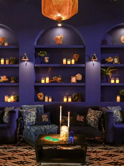 A lounge at 'Midnight Mirage', adorned with captivating cobalt hues, intriguing mystic artifacts on shelves, and the soft glow of candles, spreading magic in every corner.