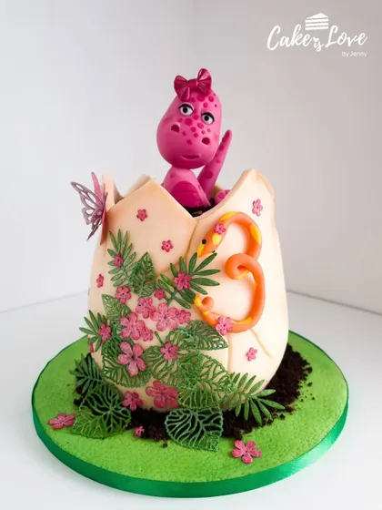 An adorable pink dinosaur hatching from an egg, decorated with tropical leaves and flowers. This is a sweet and creative take on the dinosaur theme for a third birthday.