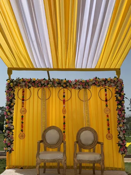 A close-up of the outdoor Haldi stage. The backdrop is decorated with floral hoops and hanging elements, framed by a border of colorful flowers.