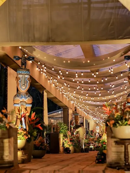 The entrance tunnel at night, with the string lights creating a warm and magical glow. The Tiki statues stand as silent guardians, welcoming guests into the celebration.