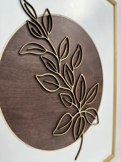 A close-up of the laser-cut leaf design. The delicate lines of the leaf are raised from the dark wood circle, creating a subtle 3D effect.