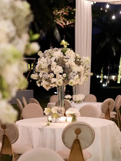 The night view of the tables, where the white floral arrangements are beautifully illuminated by candlelight and overhead bistro lights. The scene is pure romance and elegance.